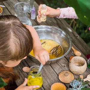 May include: A child is adding yellow liquid to a bowl of yellow liquid and brown sticks. The bowl is on a wooden table with a small jar of yellow liquid, a wooden spoon, and a wooden lid.