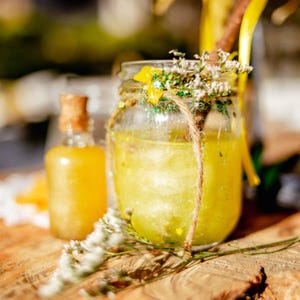 May include: A clear glass jar with a yellow liquid inside, decorated with twine and dried flowers. The jar is sitting on a wooden surface with a smaller bottle of yellow liquid next to it.