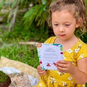 May include: A young girl holds a card with a colorful floral design and the text "I am a Powerful Shining Light. I can rise and shine everyday, like the Sun and the Moon in the sky. With my heart full of light, I make this world a happy and beautiful place. I am a bright and colorful being of light."