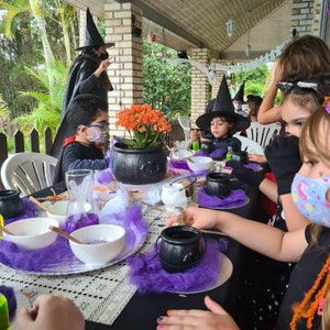 May include: A Halloween-themed party table setting with purple tablecloths, black cauldrons, and a variety of potions. Children are dressed as witches and wizards, enjoying a magical feast.