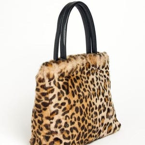 May include: A tote bag featuring a tan and brown leopard print faux fur exterior. The bag has two black handles attached to the top edge. The body of the bag has a rectangular shape with a soft, textured surface and a distinct animal pattern.