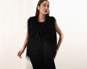 May include: A black faux fur vest with a shaggy texture. The sleeveless vest has a round neckline and a straight hemline. The vest is worn over a black outfit. The background is a plain white wall.