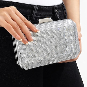May include: A rectangular silver clutch covered in small, sparkling rhinestones. The clutch has a rectangular shape with a silver clasp. The bag is held by a person wearing black jeans and a white top.