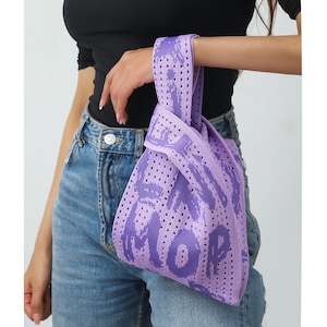 May include: A lavender tote bag with a perforated design and the word "MORE" in a graffiti-style font. The bag has a handle and is held by a person wearing a black top and blue jeans. The bag is a stylish accessory.