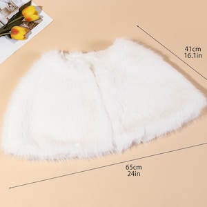 May include: A white faux fur cape, 24 inches long and 16.1 inches wide. The cape is laid flat on a light brown surface. A magazine and yellow tulips are in the upper left corner.
