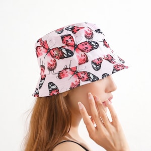 May include: A pink bucket hat featuring a repeating pattern of red and black butterflies. The hat is made of a soft fabric and has a classic, casual style. This hat is a fashionable accessory for sunny days.