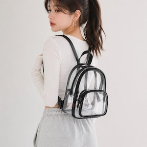 May include: A clear, transparent backpack with black trim and straps. The backpack has a small front pocket and a top handle. The bag is worn over the shoulders.