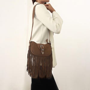 May include: A brown suede crossbody bag with long fringe and a buckle closure. The bag has a long strap and is worn over the shoulder. The bag is a medium size and is a classic style.