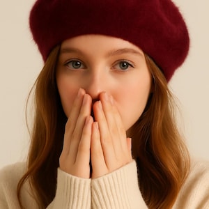 May include: A burgundy beret made of soft material. The beret is round and sits atop the head. The person in the image is wearing a cream-colored sweater.