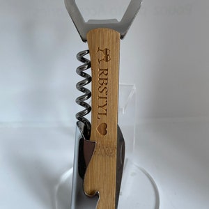 Könnte beinhalten: Ein Flaschenöffner aus Holz mit einem Korkenzieher, einem Flaschenöffner und einem Messer. Der Griff ist mit dem Text "RBSTY" und einem Herzen graviert.