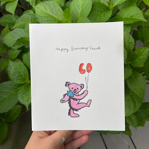 May include: A white greeting card with the text "Happy Birthday Frank" in black script. A pink bear illustration holds two red balloons with the number "60". The card is held against a backdrop of green leaves.