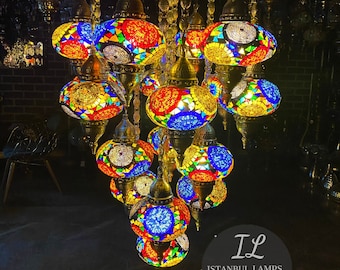 Könnte beinhalten: Ein mehrstufiger Kronleuchter mit runden, bunten Mosaikglas-Kugeln. Der Kronleuchter ist aus Messing und hat eine goldene Oberfläche. Die Kugeln sind in Blautönen, Rot, Gelb und Grün gehalten. Der Kronleuchter hängt von der Decke und ist beleuchtet. Der Text "IL ISTANBUL LAMPS LIGHTING FACTORY" ist in der unteren rechten Ecke des Bildes sichtbar.