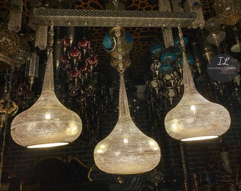 May include: Three silver pendant lights with a geometric design hang from a silver bar. The lights are illuminated and cast a warm glow. The lights are surrounded by other decorative lights in a shop.