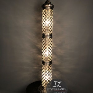 May include: A tall, silver floor lamp with a geometric patterned glass shade. The lamp has a decorative finial on top and sits on a round, gold base. The lamp is lit and casting a warm glow. The image also includes the text "IL Istanbul Lamps Lighting Factory".