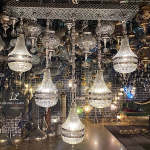 May include: A shop filled with ornate, hanging glass and metal chandeliers. The chandeliers are in various shapes and sizes, with some featuring intricate designs. The shop is decorated with a dark brick wall and a green velvet sofa. The sign on the wall reads "IL Istanbul Lamps Lighting Gallery".