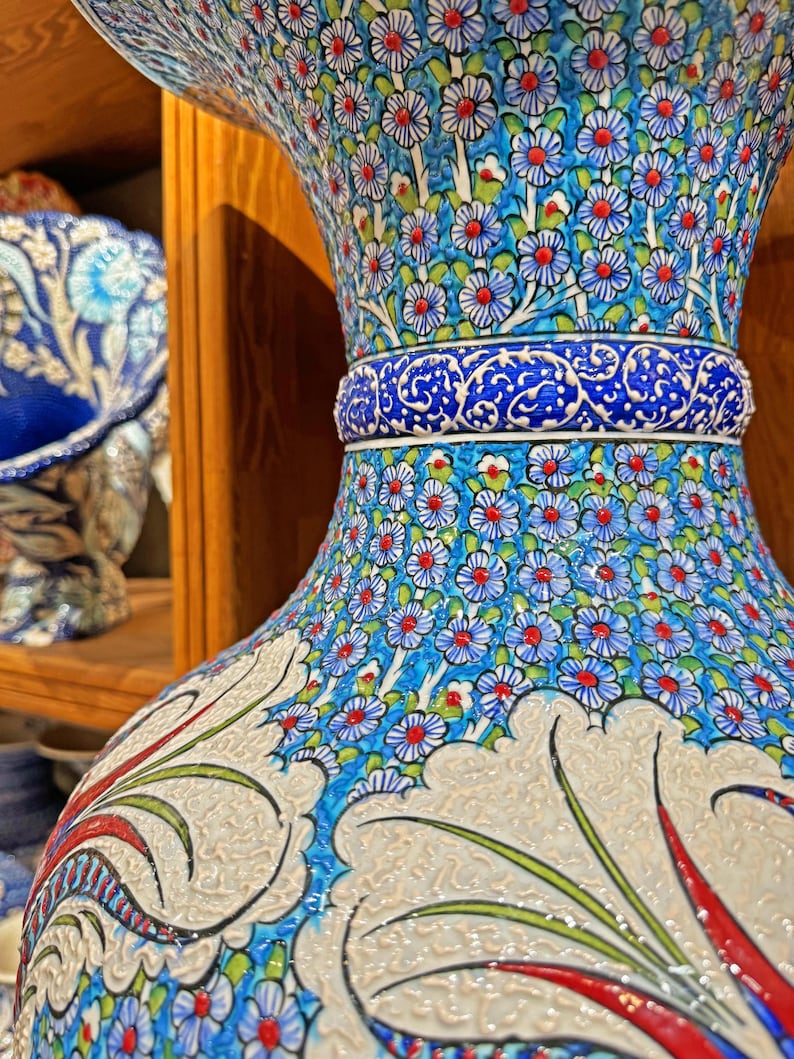 May include: A large, blue and white ceramic vase with intricate floral designs. The vase has a wide mouth and a narrow base, and is decorated with red, green, and white accents. The vase is a traditional Turkish design.