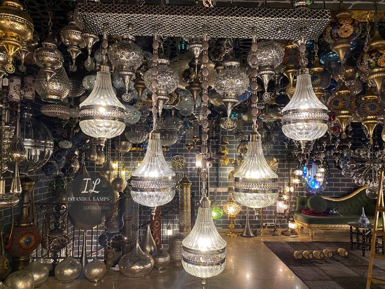 May include: A shop filled with ornate, hanging glass and metal chandeliers. The chandeliers are in various shapes and sizes, with some featuring intricate designs. The shop is decorated with a dark brick wall and a green velvet sofa. The sign on the wall reads "IL Istanbul Lamps Lighting Gallery".