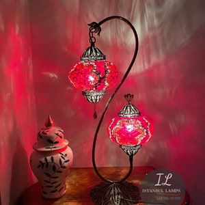 May include: A pink and white mosaic glass table lamp with two lights on a curved metal stand. The lamp is lit and casting a warm glow. The lamp is on a wooden table next to a white ceramic jar with a leaf design. The lamp has a label that reads "IL Istanbul Lamps Lighting Factory".