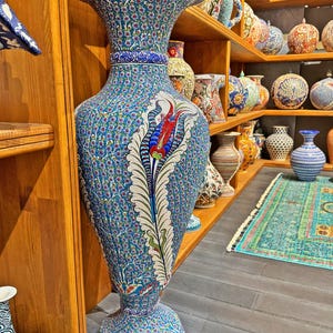 May include: A large, blue and white ceramic vase with intricate floral designs and a red tulip. The vase is on a wooden shelf with other ceramic vases.