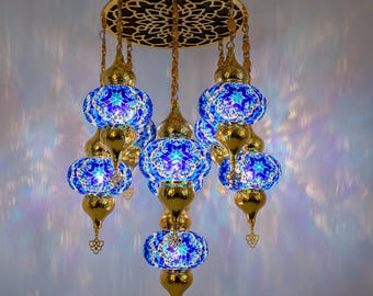 Könnte beinhalten: Ein Kronleuchter mit sieben Leuchten, bestehend aus sieben kugelförmigen Glasschirmen mit blau-weißen Mosaikmustern. Die Leuchte verfügt über eine kreisförmige Deckenplatte aus goldfarbenem Metall mit filigranen Ausschnitten und passenden Akzenten.