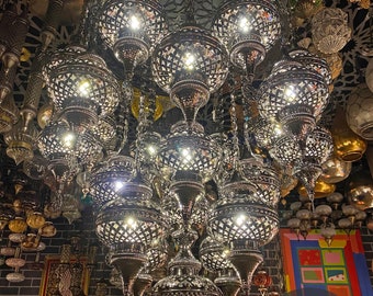 May include: A large, ornate silver chandelier with multiple tiers of round, openwork globes. The chandelier is lit and casts a warm glow. The chandelier is hanging from a ceiling with a decorative pattern. The text "ISTANBUL LAMPS LIGHTING FACTORY" is visible in the lower right corner of the image.
