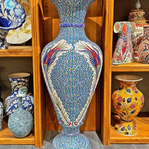 May include: A large, blue and white ceramic vase with intricate floral patterns. The vase has a wide mouth and a narrow base, and it is decorated with red and white accents. The vase is displayed on a wooden shelf.