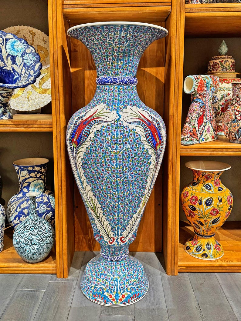 May include: A large, blue and white ceramic vase with intricate floral patterns. The vase has a wide mouth and a narrow base, and it is decorated with red and white accents. The vase is displayed on a wooden shelf.