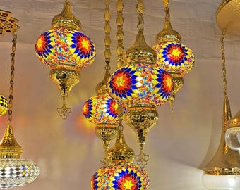 Könnte beinhalten: Ein goldfarbener Kronleuchter mit fünf bunten Mosaikglasgloben. Die Globen sind eine Mischung aus Blau, Gelb, Rot und Orange. Der Kronleuchter hat eine runde, gehämmerte Metallhaube.