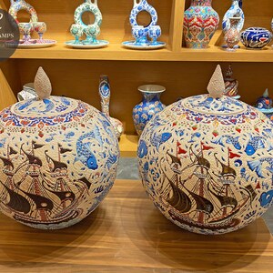 May include: Two large, round ceramic jars with lids, decorated with intricate blue, red and gold patterns. The jars feature a central design of a ship with sails, surrounded by fish and floral motifs. The jars are displayed on a wooden shelf.