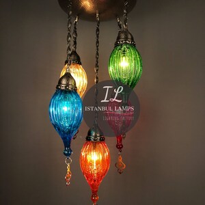 Puede incluir: Una l&aacute;mpara de ara&ntilde;a de vidrio multicolor con cinco luces. Las pantallas de vidrio son verdes, azules, naranjas y amarillas. La l&aacute;mpara de ara&ntilde;a cuelga de una fijaci&oacute;n de techo de metal. El texto "Istanbul Lamps Lighting Factory" es visible en la imagen.