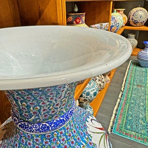 May include: A large, white ceramic vase with a wide, flared opening and a blue and white floral pattern. The vase is decorated with intricate details and is sitting on a wooden shelf.