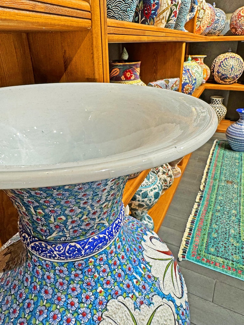 May include: A large, white ceramic vase with a wide, flared opening and a blue and white floral pattern. The vase is decorated with intricate details and is sitting on a wooden shelf.