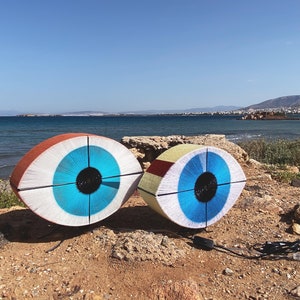 Könnte beinhalten: Zwei große, farbenfrohe, augenförmige Skulpturen aus Garn, mit blauen Iris und schwarzen Pupillen, stehen auf einem Sandstrand mit blauem Meer und einer entfernten Küstenlinie im Hintergrund.