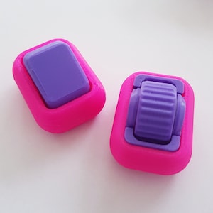 May include: Two pink plastic toggles with purple accents. The toggles are square shaped and have a small, round, purple button in the centre.