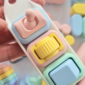 Puede incluir: Un llavero de colores pastel con tres peque&ntilde;os juguetes cuadrados. Cada juguete tiene un dise&ntilde;o diferente: un bot&oacute;n rosa, un rodillo amarillo y un bot&oacute;n azul. El llavero es blanco y tiene un anillo plateado.