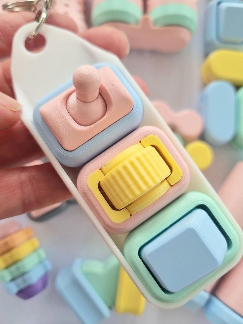 Puede incluir: Un llavero de colores pastel con tres peque&ntilde;os juguetes cuadrados. Cada juguete tiene un dise&ntilde;o diferente: un bot&oacute;n rosa, un rodillo amarillo y un bot&oacute;n azul. El llavero es blanco y tiene un anillo plateado.