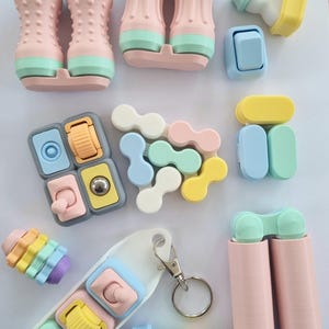Puede incluir: Una colecci&oacute;n de juguetes fidget coloridos en varias formas y dise&ntilde;os. Los juguetes presentan colores pastel rosa, azul, amarillo y verde. Algunos tienen botones, rodillos y otros elementos interactivos. Tambi&eacute;n se incluye un llavero con juguetes fidget similares.