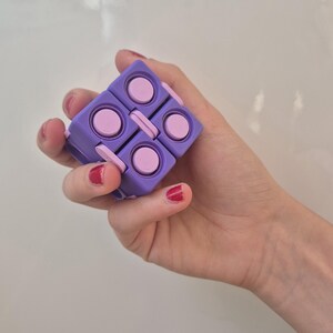 May include: A purple and light pink fidget cube toy featuring a modular design with multiple interconnected square segments. Each segment has a circular button on its surface, allowing the object to be folded and manipulated into various shapes.