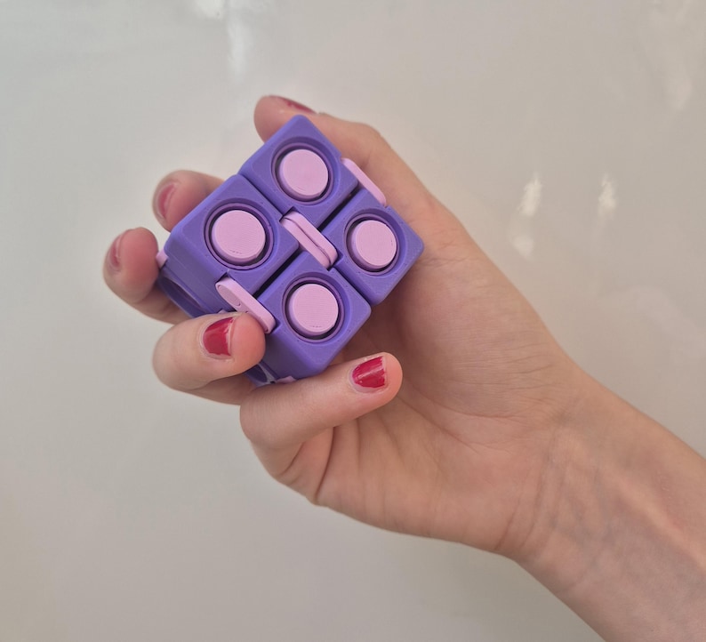 May include: A purple and light pink fidget cube toy featuring a modular design with multiple interconnected square segments. Each segment has a circular button on its surface, allowing the object to be folded and manipulated into various shapes.