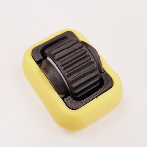 Pode incluir: Um brinquedo fidget impresso em 3D com um rolo castanho escuro texturizado, encaixado numa moldura retangular amarela. O rolo tem detalhes canelados e a moldura tem bordas arredondadas. O brinquedo foi concebido para o envolvimento tátil.