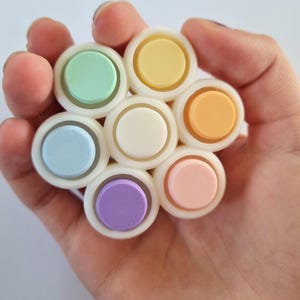 May include: A set of seven pastel-coloured round push-pop fidget toys. The toys are arranged in a circle and are made of a soft, squishy material. The colours are light blue, light green, light purple, light pink, light yellow, and two shades of white.