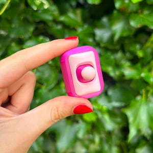 Pode incluir: Um brinquedo antiestresse retangular com uma carcaça externa rosa brilhante e um painel central rosa claro. O dispositivo inclui um botão de joystick circular central em um tom rosa claro correspondente para interação tátil.
