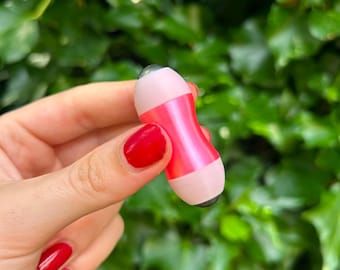 Puede incluir: Una pequeña herramienta de masaje facial manual con un cuerpo central de color rosa metálico en forma de reloj de arena, flanqueado por dos tapas de color rosa claro, cada una con una bola rodante gris oscuro en los extremos.