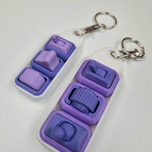 Puede incluir: Dos llaveros con bases blancas y dise&ntilde;os impresos en 3D en morado y lavanda. Cada llavero tiene tres formas diferentes, incluyendo un cuadrado, una estrella y un bot&oacute;n. Los llaveros tienen llaveros y cierres plateados.