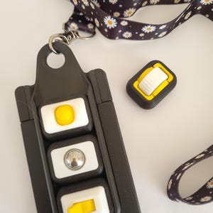 Puede incluir: Cord&oacute;n negro con un estampado de margaritas, sujeto a un soporte negro con tres botones blancos y amarillos. Tambi&eacute;n se ve un bot&oacute;n negro y amarillo separado.