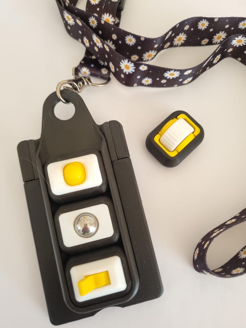 Puede incluir: Cord&oacute;n negro con un estampado de margaritas, sujeto a un soporte negro con tres botones blancos y amarillos. Tambi&eacute;n se ve un bot&oacute;n negro y amarillo separado.