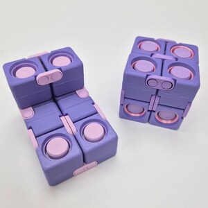 May include: Two purple and light pink fidget cubes constructed from multiple interconnected square blocks. Each block features a circular button on its surface, and the cubes are shown in both a folded, compact shape and an unfolded, elongated form.
