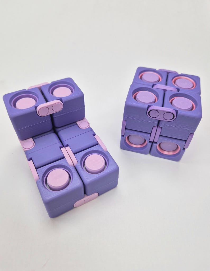 May include: Two purple and light pink fidget cubes constructed from multiple interconnected square blocks. Each block features a circular button on its surface, and the cubes are shown in both a folded, compact shape and an unfolded, elongated form.