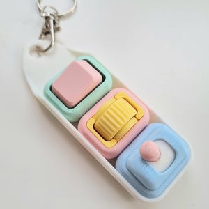 Puede incluir: Un llavero de colores pastel con tres colgantes cuadrados. Los colgantes son rosas, amarillos y azules, cada uno con un dise&ntilde;o &uacute;nico. El llavero incluye un cierre plateado y una base oblonga blanca.