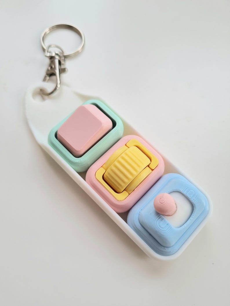 Puede incluir: Un llavero de colores pastel con tres colgantes cuadrados. Los colgantes son rosas, amarillos y azules, cada uno con un dise&ntilde;o &uacute;nico. El llavero incluye un cierre plateado y una base oblonga blanca.